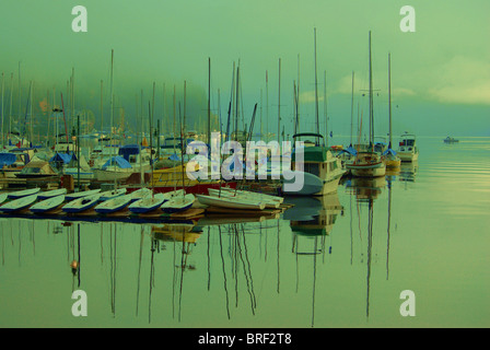 Small marina on foggy morning with masts of sailboats reflecting on calm water surface. Stock Photo