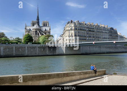 Two females sitting on a wall at the edge of the River Seine with Notre Dame in the background, and residential buildings beside Stock Photo