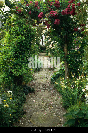 Circular paving stones and cobble paving on path through arched trellis with red climbing rose in country garden Stock Photo