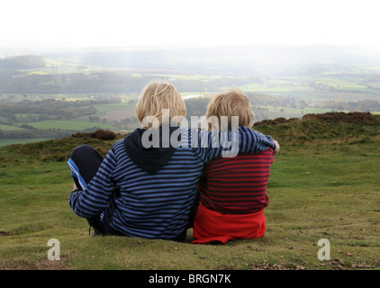 Two blond-haired brothers sitting on hillside one is comforting the other with his arm around his shoulder after some sad news Stock Photo