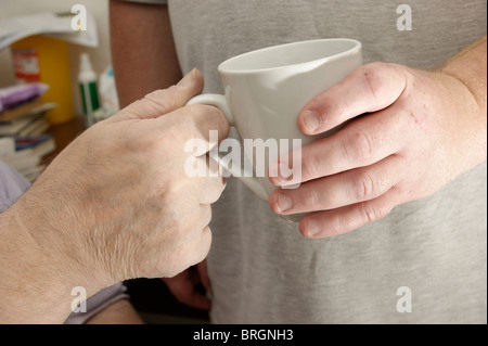 male carer (son) giving a cup of tea / coffee to a disabled elderly man Stock Photo
