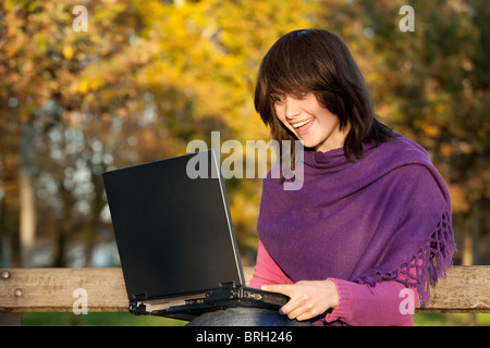 young woman beauty laughing using computer seated on park bench in warm sunlight Stock Photo