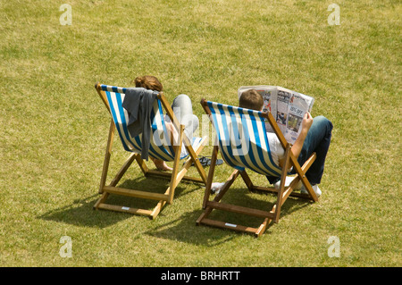 Horizontal aerial back view of a couple, man and a woman, relaxing and sunbathing in a park sitting on striped wooden deckchairs Stock Photo