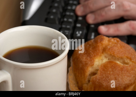 cup of black coffee and a muffin in front of a mans hand typing on a computer keyboard Stock Photo