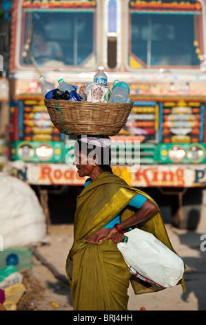 Old Indian woman carrying a basket of bottles on her head in front of an indian lorry. Andhra Pradesh, India Stock Photo