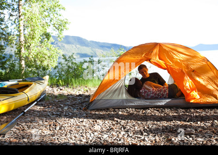 A young couple camping talk in their tent next to kayaks with lake in the background in Idaho. Stock Photo