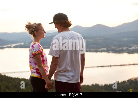 A young adult couple hold hands and smile at sunset overlooking a large lake. Stock Photo