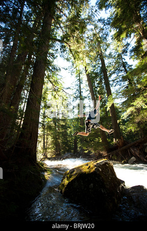 A young man hiking jumps off a rock next to a river in the forest. Stock Photo