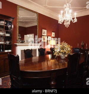 Lighted chandelier above antique table and chairs in traditional red dining room with large gilt mirror above fireplace Stock Photo