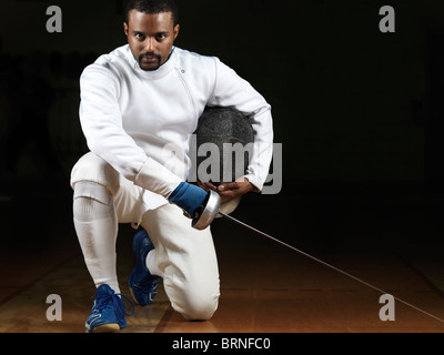 Portrait of a fencer wearing fencing uniform and holding an epee in a gym on black background Stock Photo