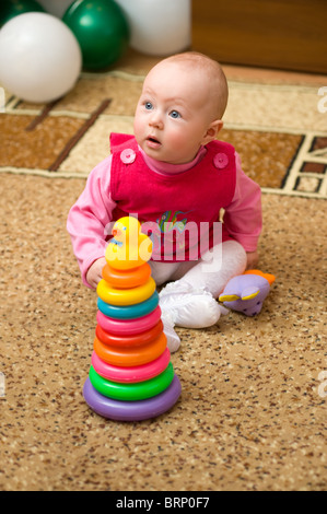 little european eight months old girl in red dress sits on carpet with toy pyramid Stock Photo