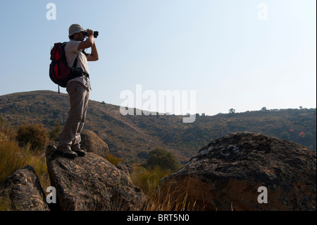 Young man with backpack looking through binoculars on top of a rock against the sky Stock Photo