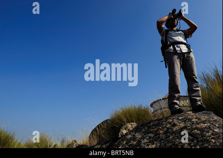 Young man with backpack looking through binoculars on top of a rock against the sky Stock Photo