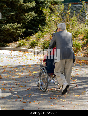 Old man pushing a lady in a wheel chair down a street through late afternoon shadows. Back view of subjects no faces shown. Stock Photo