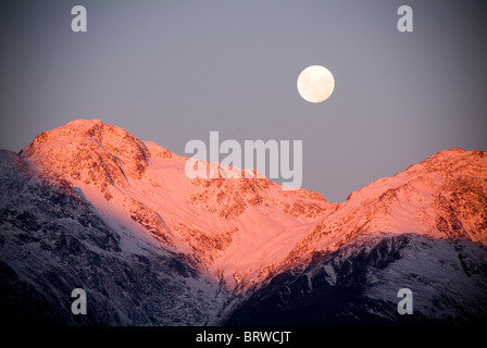 Moon rises over snow capped mountains at twilight Stock Photo