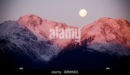 Moon rises over snow capped mountains at twilight Stock Photo