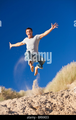 A happy and excited young man jumping high with arms outstretched on a sunny sandy beach with blue sky Stock Photo