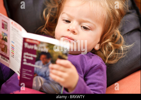 A toddler girl relaxing on a sofa reads a Turkish language study book. Stock Photo