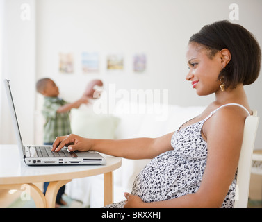 Pregnant Black mother using laptop while son plays with football Stock Photo