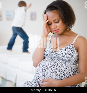 Pregnant Black mother sitting with head in hands while son jumps on bed Stock Photo