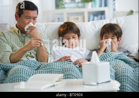 Sick father and sons blowing noses and checking temperature on living room sofa Stock Photo