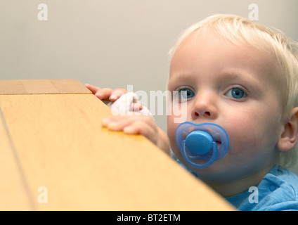 Toddler looking over a table with dummy in mouth Stock Photo