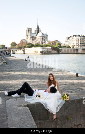 pretty Asian bride sits gacefully with groom's head in her lap on Quai de la Tournelle against backdrop of Notre Dame de Paris Stock Photo