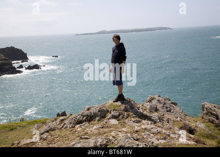 Teenage boy stands in strong wind on rocky headland above sea Stock Photo