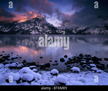 A pink sunrise reflection on a frozen Jenny Lake shows first light through a winter storm in Grand Teton National Park, Wyoming. Stock Photo