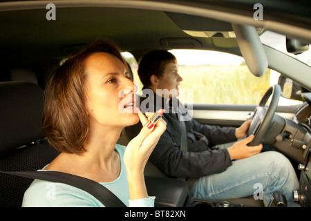 Young woman applying red lipstick makeup in car passenger seat close up female beauty cosmetics while boyfriend drives road trip travel journey Stock Photo