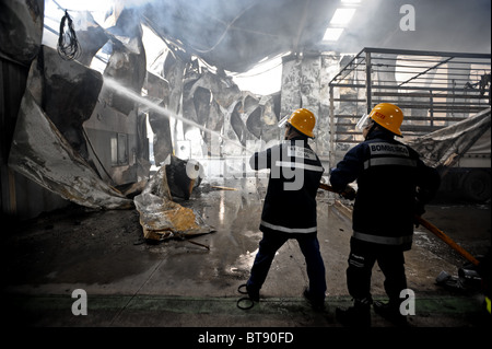 Two firemen help each other while fighting a factory fire Stock Photo