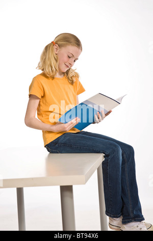 Blonde girl with pigtails sitting on a table and reading a schoolbook Stock Photo