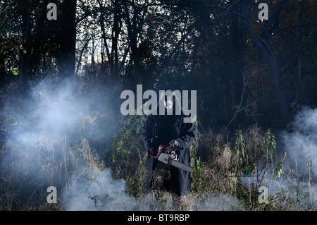 Glaring pale evil man in black coat with a chainsaw in a misty forest in the Fall at Halloween Stock Photo