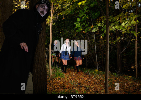 Three happy schoolgirls walking on a path in the woods with a sinister man in black cloak waiting to ambush them in the isolated forest Stock Photo