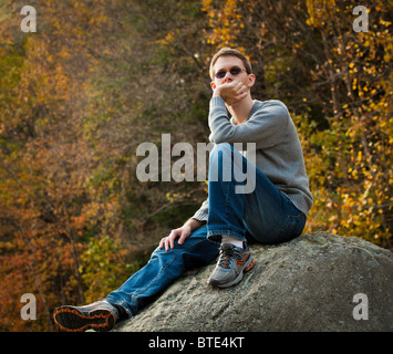 Young male hiker in jeans pondering while sitting on a boulder in front of fall leaves Stock Photo