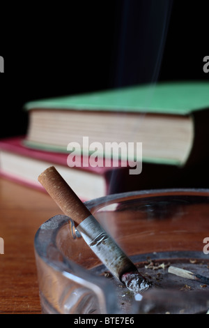 Burning cigarette with smoke in an ash tray with old books in the background Stock Photo