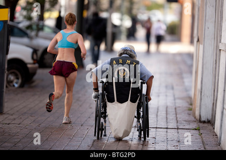 A man in a wheelchair wearing an vest emblazoned with Army insignia pushes himself up the sidewalk as woman jogs by Stock Photo