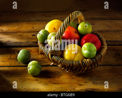 Mixed red, yellow and green plums in a basket on a wooden table Stock Photo