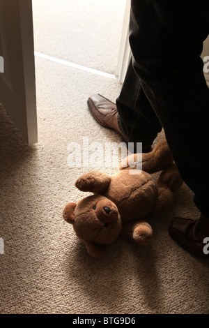 Man (feet and legs only) wearing brown shoes stepping over a teddy bear lying on the floor by an open door. Stock Photo