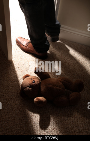 Man (feet and legs only) wearing brown shoes stepping over a teddy bear lying on the floor by an open door. Stock Photo