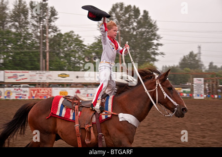 This young 6 year old boy is trick riding this beautiful brown horse in a rodeo in the rain. His hat is off greeting the crowd. Stock Photo