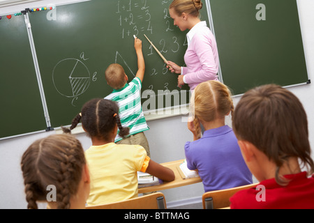 Photo of elementary student doing sums on blackboard and looking at it while his teacher helping him Stock Photo