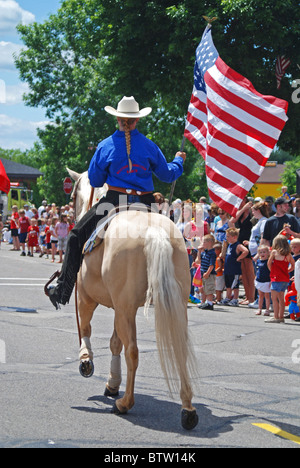 Cowgirl rides her white horse in the July 4th parade in rural America carrying a USA flag as she walks the horse through crowds Stock Photo