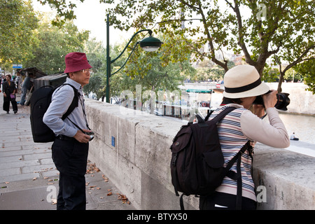 middle aged Japanese tourist couple wearing sunhats knapsacks & cameras with husband watching wife photograph Seine river view Stock Photo