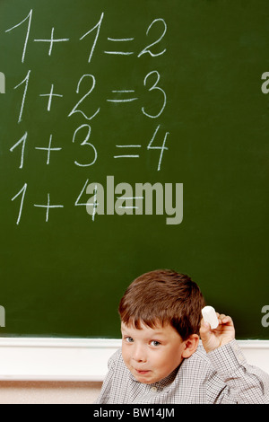 Portrait of curious schoolchild standing at blackboard with written sums on it and looking at camera Stock Photo