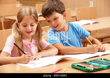 Portrait of two schoolchildren sitting by one desk during drawing lesson while boy looking at his mate’s copybook Stock Photo