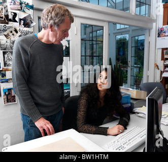 Two Work Colleagues Chatting In office Asian Woman Sitting by Computer And Man Standing Next to Her Stock Photo