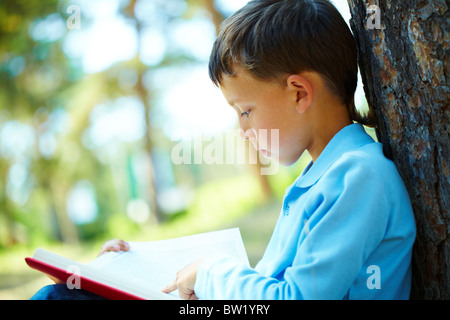 Portrait of smart boy sitting by tree trunk in the park and reading book Stock Photo