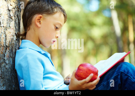 Portrait of smart boy sitting by tree trunk in the park and reading book Stock Photo
