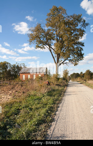 Abandoned on-room schoolhouse on a rural, gravel road in Indiana with a large tree, bright blue sky and clouds vertical Stock Photo